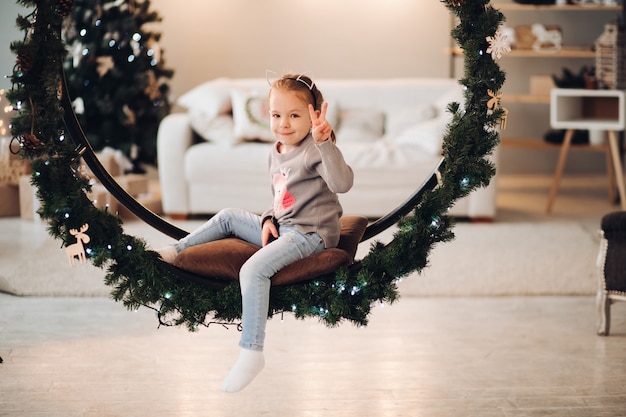Pretty child sitting on Christmas swing. Christmas tree.
