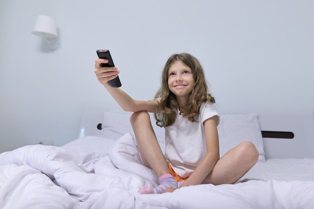 Довольно ребенок женщина блондинка утром, сидя в белой кровати с пультом дистанционного управления телевизором