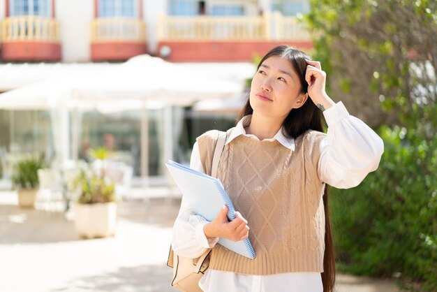 Pretty Chinese student woman at outdoors having doubts and with confuse face expression