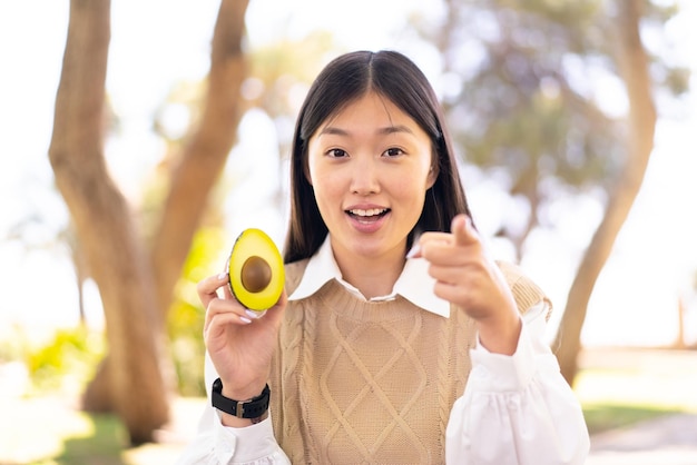 Pretty Chinese woman holding an avocado at outdoors surprised and pointing front