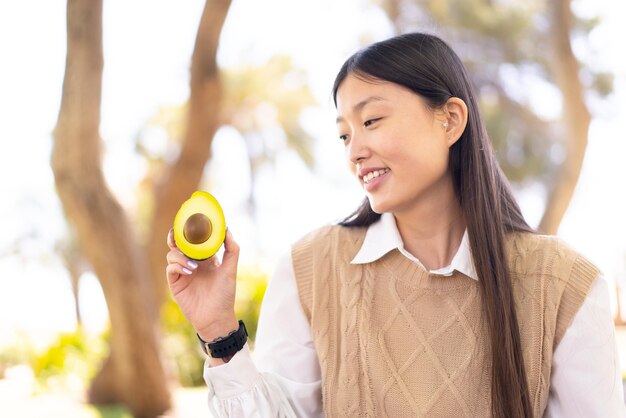 Pretty Chinese woman holding an avocado at outdoors with happy expression