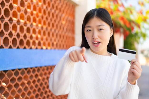 Pretty Chinese woman holding a credit card at outdoors surprised and pointing front