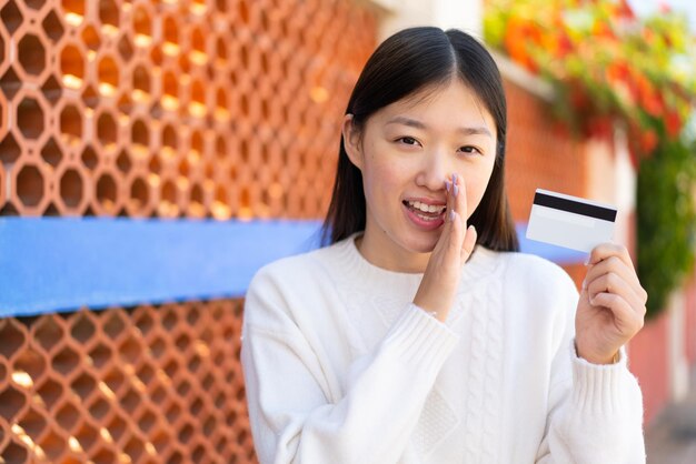 Pretty Chinese woman holding a credit card at outdoors whispering something