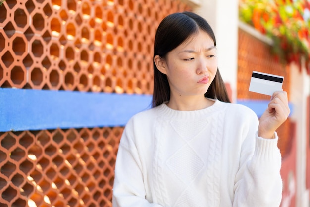 Pretty Chinese woman holding a credit card at outdoors with sad expression