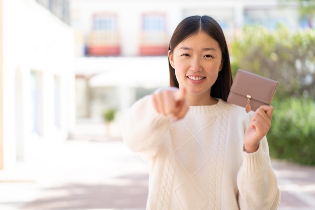 屋外で財布を持ったかわいい中国人女性が自信に満ちた表情であなたに指を向ける