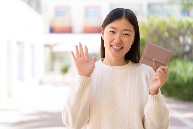 Pretty Chinese woman holding a wallet at outdoors saluting with hand with happy expression