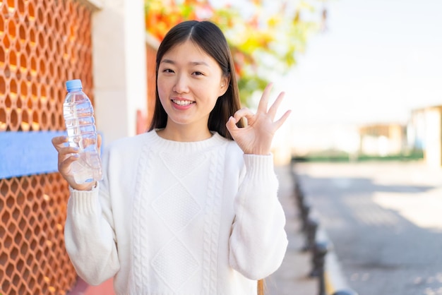 Красивая китаянка с бутылкой воды на улице показывает пальцами знак "ок"