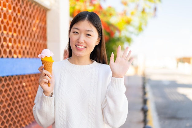 야외에서 코넷 아이스크림을 들고 행복한 표정으로 손으로 경례하는 예쁜 중국 여성