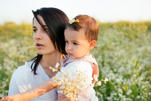 Pretty close up portrait of mother enbraces a daughter toddler baby surprised and looking at one side with flowers in hand on field nature background Motherhood and childhood