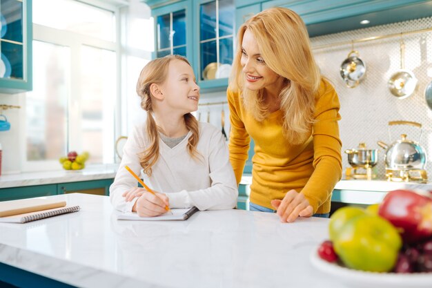 Pretty content fair-haired girl smiling and holding a pencil while looking at her mother standing behind her