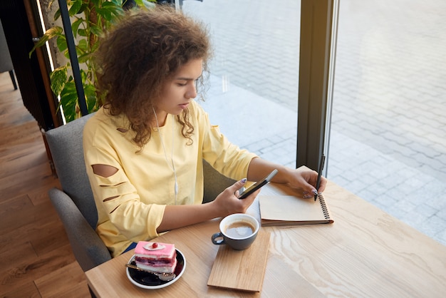 かなり巻き毛の少女がメモを書いて、カフェでコーヒーを飲みます。