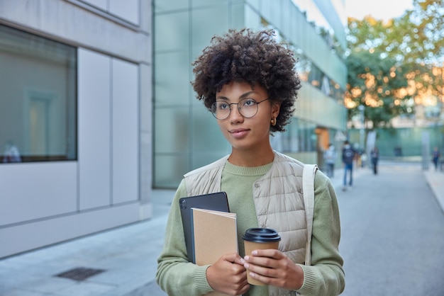 Pretty curly haired woman holds digital tablet notepad paper cup of coffee focused into distance wears round spectacles casual outfit strolls outdoors during sunny day Student on way to university