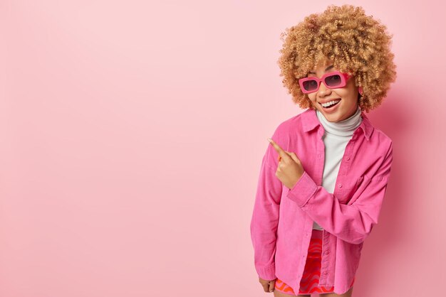 Pretty curly haired woman smiles gladfully wears sunglasses and\
shirt points index finger on blank space advertises product\
isolated over pink background click on this link and follow this\
direction