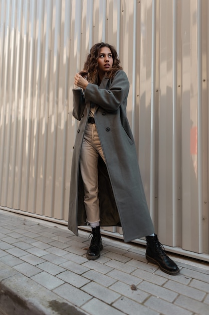 Pretty curly model woman in vintage fashion long coat and boots stands near a metal wall on the street. Urban casual modern style and female beauty