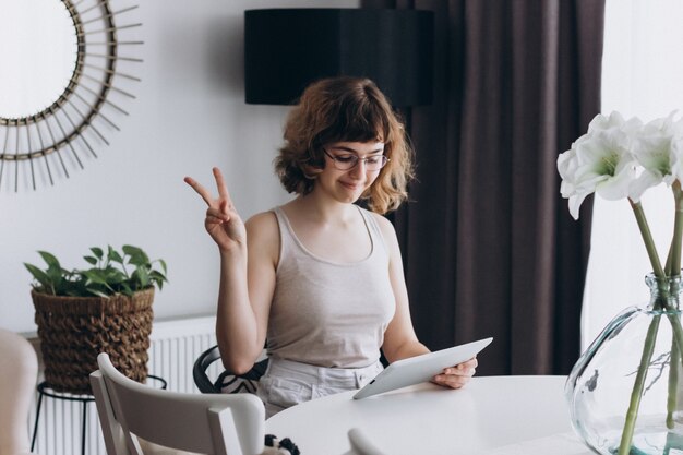 Pretty curly smiling young girl talking by phone sitting at the table at home. Video chatting with friends