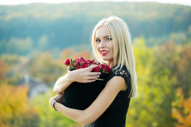 Pretty cute girl or beautiful blond woman in sexy black dress with red rose flowers in box outdoor on natural background