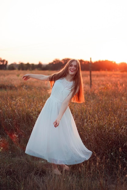 Pretty cute girl is circling in a white dress on the field. Sun rays. Orange sunset. Concept. A woma