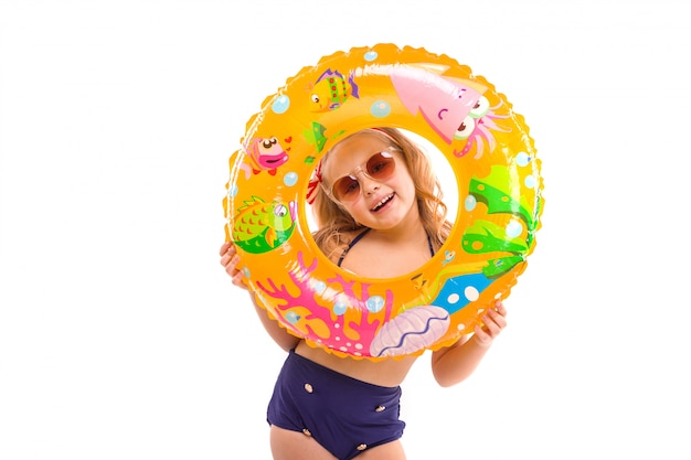 Pretty cute girl in red striped bikini, blue bottoms, sunglasses and pink wreath stand stand with rubber ring in hand