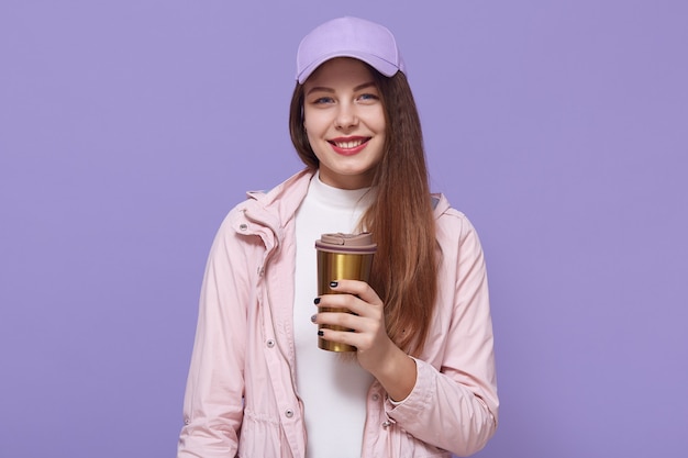 Довольно темноволосая европейская женщина с длинными волосами, носит куртку и бейсболку, держит кофе в термокружке, модели над лиловой стеной, смотрит, улыбаясь в камеру.