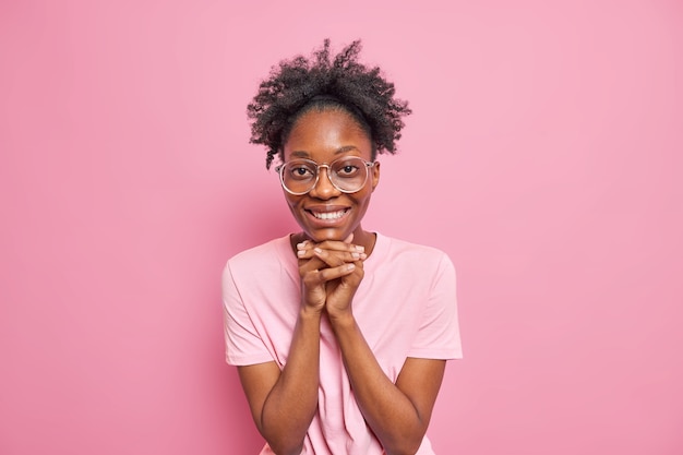 Pretty dark skinned woman keeps hands under chin smiles positively looks directly at camera