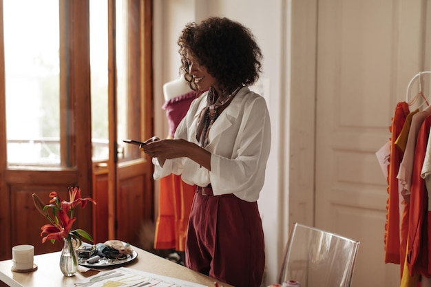 Pretty darkskinned curly lady in burgundy pants and white blouse holds phone and smiles Beautiful fashion designer poses in office