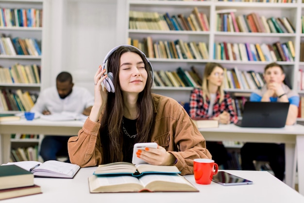 Ragazza di 25 anni piuttosto sognante in camicia casual che indossa le cuffie e che gode della musica preferita durante la pausa dopo un duro studio