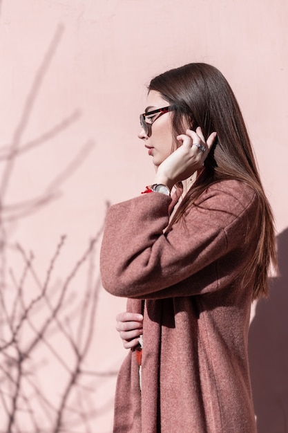 Pretty elegant young woman fashion model with gorgeous long hair in fashionable coat in trendy sunglasses posing near vintage pink wall on bright sunny day. Stylish girl in spring wear posing in sun.