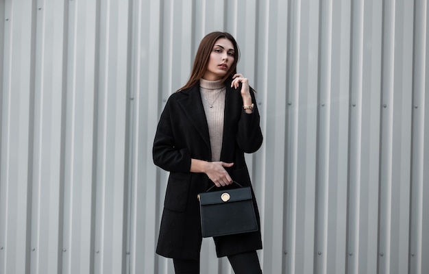 Pretty elegant young woman fashion model with long hair in black spring trendy coat with fashionable leather black handbag stands near a vintage metal wall in the city. Cute luxurious girl. Urban lady