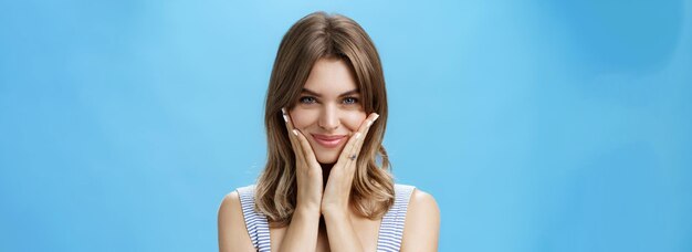 Pretty european girl pressing palms to cheeks and smiling happily while looking at camera as if person