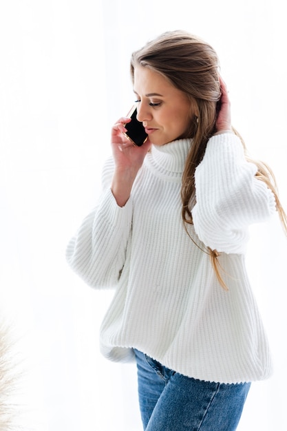 Pretty european woman in casual white sweater sit at home by window with phone, positive talking, having conversation