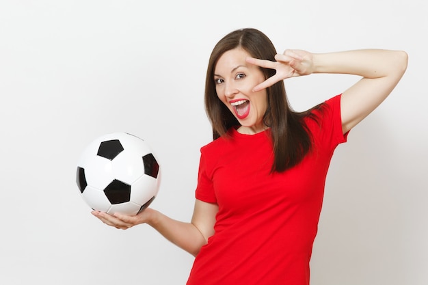 Pretty european young woman, football fan or player in red\
uniform holding classic soccer ball, fingers near eye isolated on\
white background. sport, play football, health, healthy lifestyle\
concept.