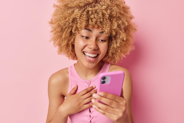 Pretty excited woman focused at smartphone screen laughs at\
something funny doesnt find her sincere feelings keeps hand on\
chest wears t shirt isolated over pink background emotions\
concept