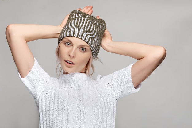 Foto modello di moda grazioso in maglione bianco e cappello lavorato a maglia tiene le mani dietro la testa