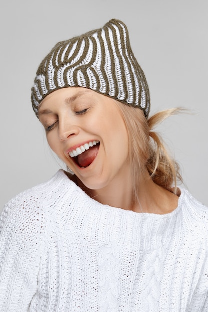 Pretty fashion model in white sweater and knitted hat laughing with wide open mouth