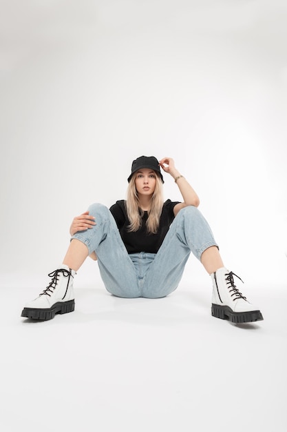 Pretty fashion young woman model with cool Panama hat in fashionable blue jeans white stylish leather shoes and tshirt sits on a white background Cool beauty girl with trendy outwear