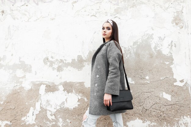 Pretty fashionable young woman with stylish black bag in trendy gray coat with glamour bandana rests near white vintage wall outdoors. Lovely girl in fashion spring youth clothes posing in street.