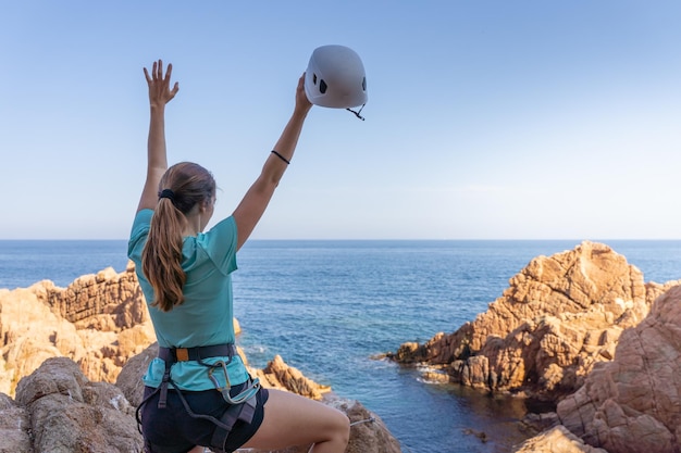 Pretty female climber celebrating strong success with arms raised on rocks over the sea with rope and helmet