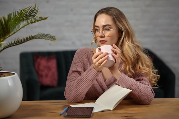 Pretty female in glasses, pink cardigan. She is holding cup, sitting at wooden table with a tablet, notebook, blue pen and flower in a pot on it. Student, blogger. Work or education concept. Close-up