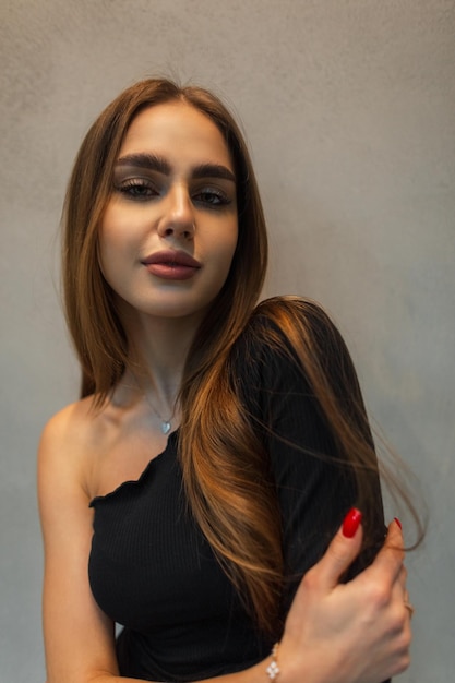 Pretty female indoors portrait of beautiful woman with fresh face in black top stands and poses near a gray wall