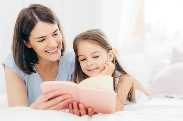 Pretty female mother and her small daughter read interesting\
book in bedroom lie on comfortabled bed during morning lovely mum\
reads fairty tale for her little female kid who is very\
curious