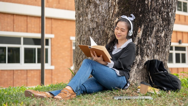 Photo a pretty female student is listening to music and reading a book relaxing under the tree in a park