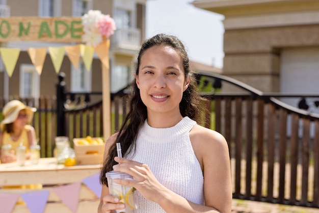 Pretty female with dark long hair having glass of fresh cool homemade lemonade that she bought by stall near by on hot summer day