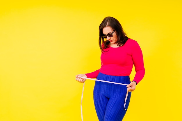 Pretty female with excess weight in sporty top measuring waist over white yellow background