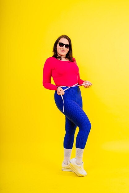 Photo pretty female with excess weight in sporty top measuring waist over white yellow background