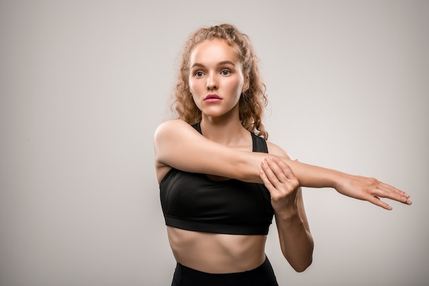 Pretty fit sportswoman in black tracksuit stretching right arm in front of herself while training on grey in gym