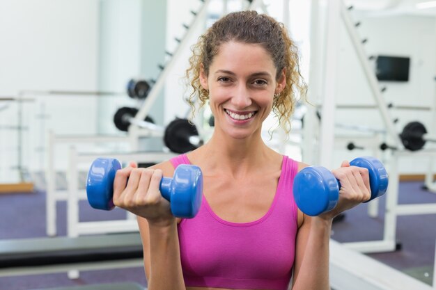 Pretty fit woman lifting blue dumbbells smiling at camera