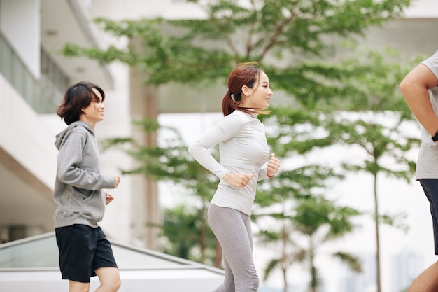 Pretty fit young Asian woman in comfy sports clothes jogging outdoors with group of friends