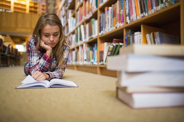 Pretty focused student lying on library floor reading book