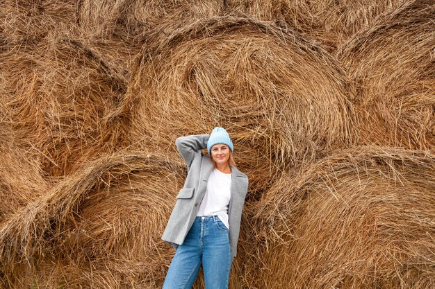 Giovane donna dai capelli scuri abbastanza fresca che cammina all'aperto al sole sullo sfondo di mucchi di fieno e indossa un cappotto, cappello da lavoro a maglia, jeans. concetto di vacanze autunnali al villaggio e stile dal vivo