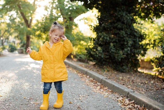 Фото Красивая смешная девочка носит желтый плащ и резиновые сапоги гуляет в парке по упавшим листьям